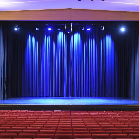 Leere Theaterbühne mit blau beleuchtetem Vorhang und roten Sitzreihen im Vordergrund.