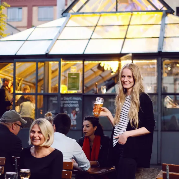 Lächelnde Menschen sitzen und stehen in einem sonnigen Biergarten vor einem Glashaus.