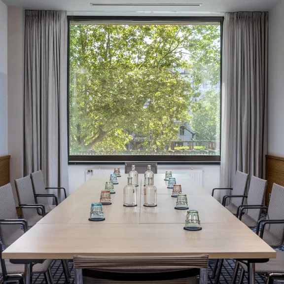 Moderner Konferenzraum mit großem Tisch, Stühlen und Blick auf grüne Bäume durchs Fenster.