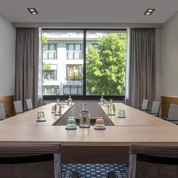 Moderner Konferenzraum mit U-förmigem Tisch, Wasserflaschen und Blick auf grüne Bäume.