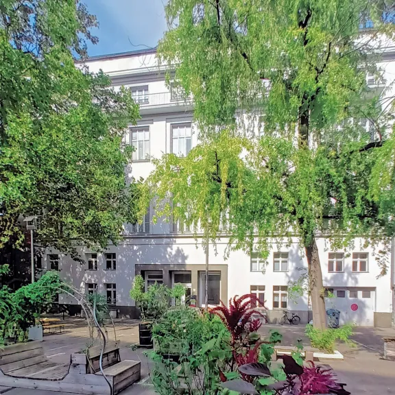 Grüner Innenhof mit Bäumen, Bänken und weißer Hausfassade im Hintergrund.