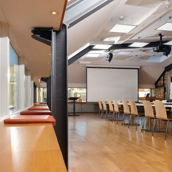 Heller Konferenzraum mit langen Tischen, Holzstühlen und großer Fensterfront