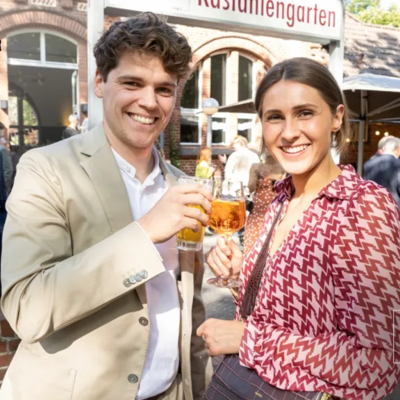 Zwei lächelnde Personen stoßen im Freien mit Getränken an, im Hintergrund stehen weitere Gäste.