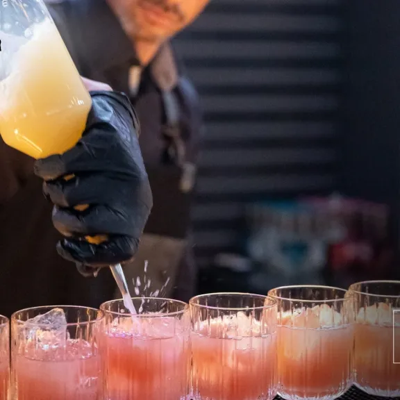 Barkeeper gießt gelbes Getränk in mehrere Gläser mit rosa Flüssigkeit.