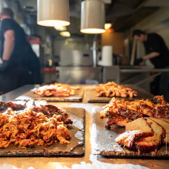 Verschiedene Portionen Fleisch auf Tabletts in einer Restaurantküche, zwei Köche im Hintergrund.