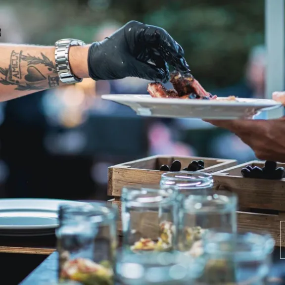 Tätowierter Arm mit schwarzem Handschuh reicht einen Teller in einem Restaurant.