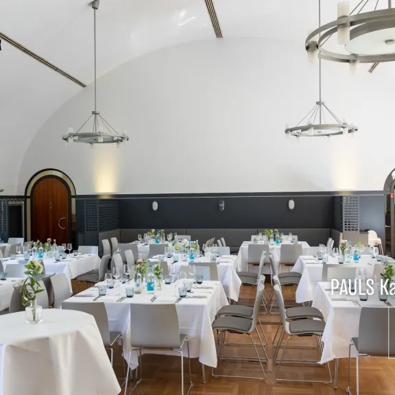 Festlich gedeckter Saal mit runden Tischen, weißen Tischdecken und Blumenvasen.