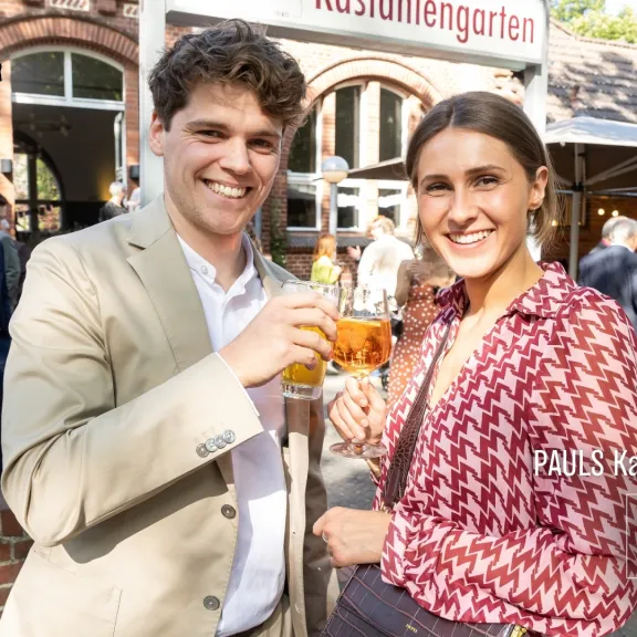 Zwei junge Menschen stoßen lachend mit Getränken in einem sonnigen Biergarten an.