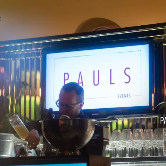 Barkeeper schenkt Getränk vor beleuchtetem PAULS-Schriftzug ein.