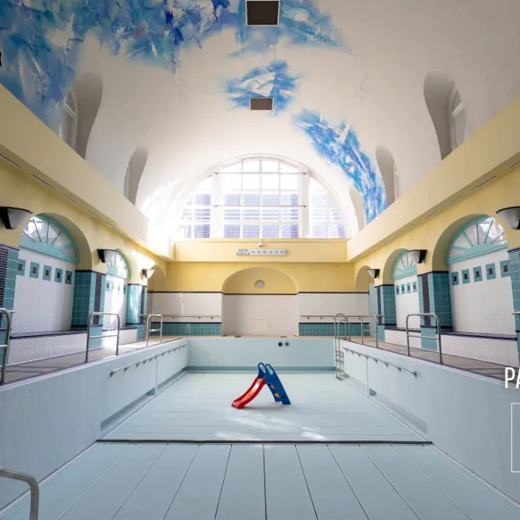 Leeres Schwimmbad mit bemalter Gewölbedecke und Licht durch ein großes Fenster.