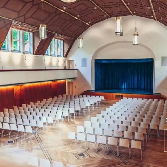 Großer, heller Saal mit Reihen weißer Stühle und Bühne mit blauem Vorhang.