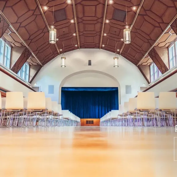 Leerer Saal mit Reihen von Stühlen und einer Bühne im Hintergrund.