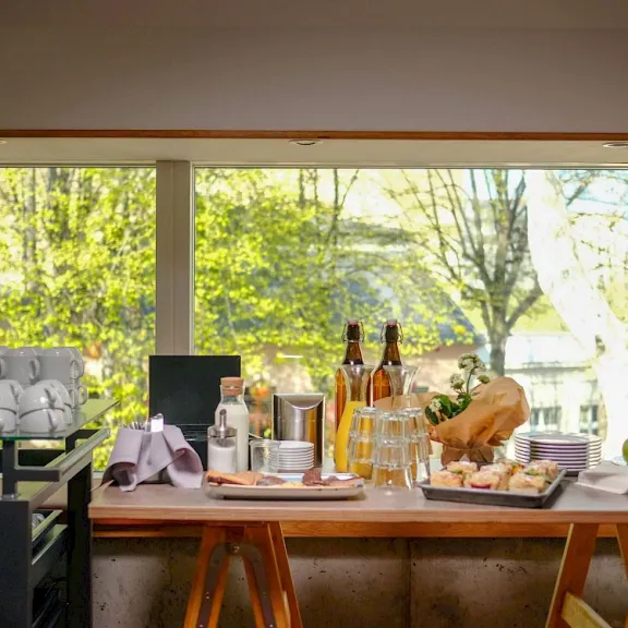 Holztisch gedeckt mit Getränken, Brot, Äpfeln und Gläsern vor großem Fenster mit Gartenblick.