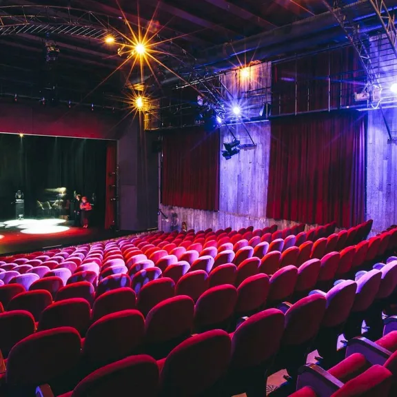 Leerer Theatersaal mit roten Stühlen, beleuchtete Bühne im Hintergrund.