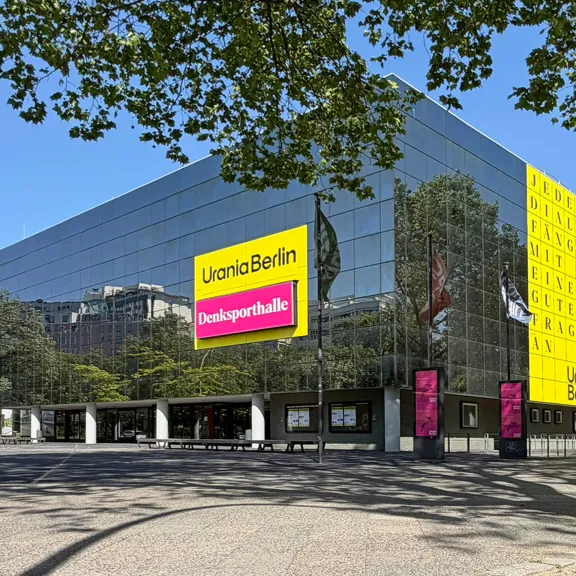 Urania mit Humboldt Saal in Berlin mit gelben und pinken Bannern, umgeben von Bäumen.
