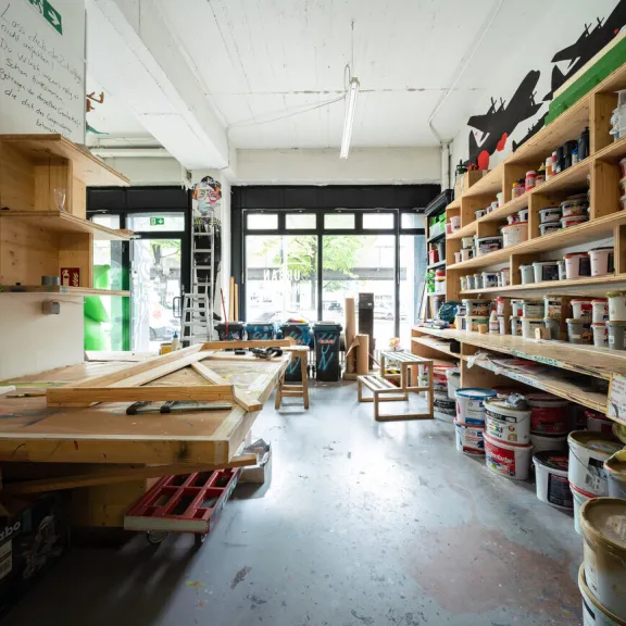 Helles Atelier mit Regalen voller Farbdosen und großem Arbeitstisch.