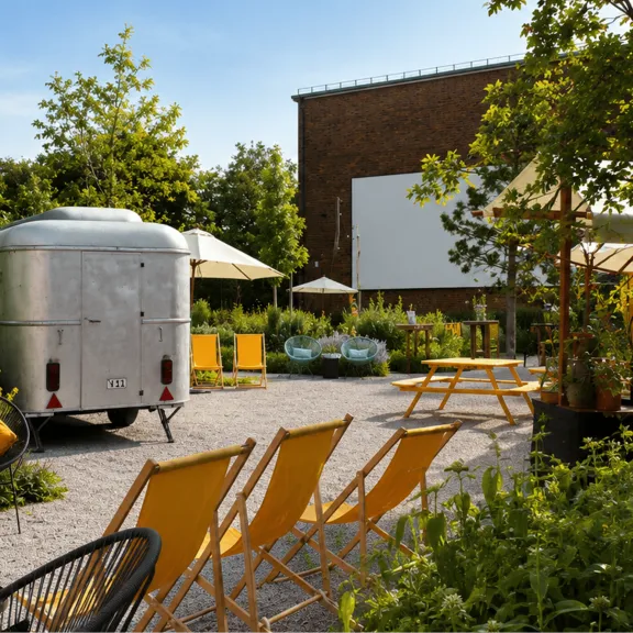 Gemütlicher Garten mit gelben Liegestühlen, Wohnwagen und viel Grün an einem sonnigen Tag.