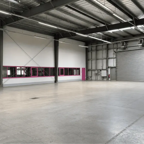 Leere Industriehalle mit Fenstern und großem Rolltor, Betonboden, Stahlträgern.