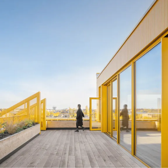 Mensch steht auf moderner Dachterrasse mit gelbem Geländer und Glasfassade, klarer Himmel.