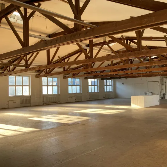 Großer, leerer Raum mit Holzbalken-Decke und Sonnenlicht durch große Fenster.