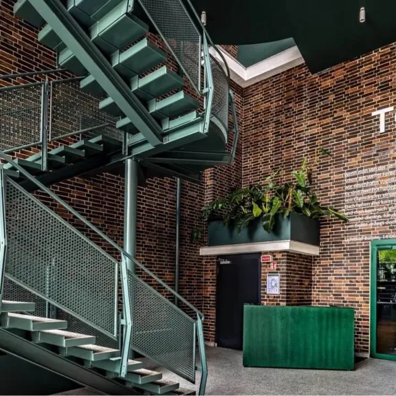 Modernes Foyer mit Backsteinwänden, grünem Tresen und Metalltreppe, Schild „TON 1“.