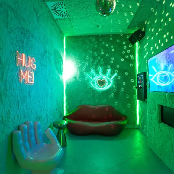Grüner Raum mit Lippen-Sofa, Handstuhl, Neonlichtern und Wandleuchten in Augenform.