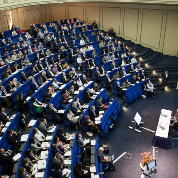 Hörsaal mit vielen Menschen auf blauen Sitzen, zwei Vortragende sitzen an Tisch