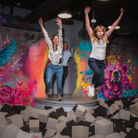 Zwei Frauen springen in einen Schaumstoffpool vor bunter Graffiti-Wand.