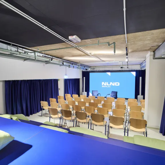 Moderner Konferenzraum mit Holzstühlen, blauen Vorhängen und großer Leinwand vorne.