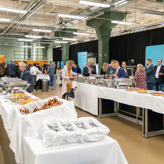 Menschen bedienen sich an einem Buffet bei einer Veranstaltung in einem großen, hellen Raum.