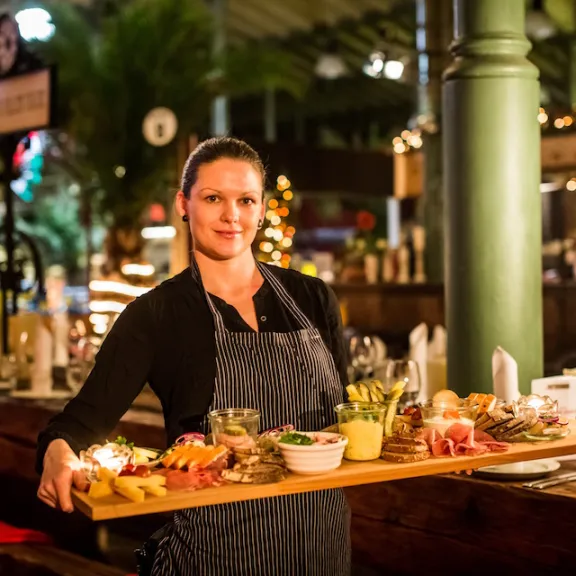 Kellnerin trägt ein großes Holzbrett mit verschiedenen Speisen in festlich dekoriertem Restaurant.