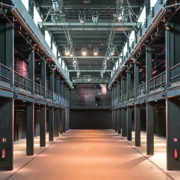 Große, leere Industriehalle mit Metallbalken und hoher Decke, Licht von oben.