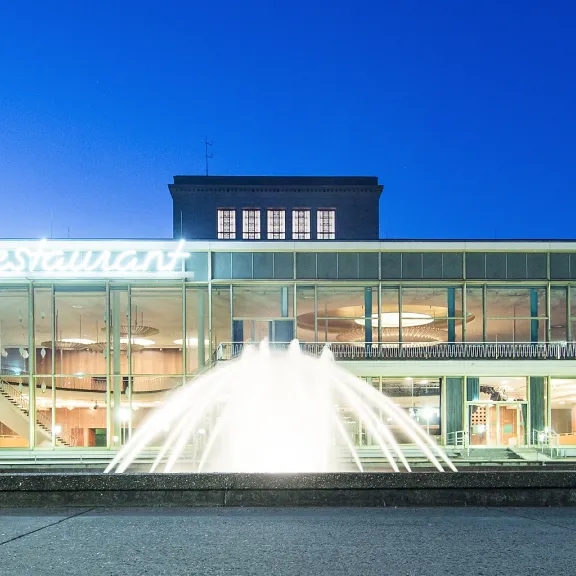 Modernes Gebäude mit großer Glasfront und beleuchtetem Springbrunnen am Abend.