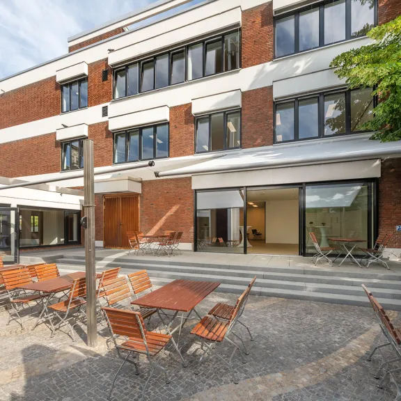 Terrasse mit leeren Holztischen und -stühlen vor modernem Gebäude bei Sonnenschein.