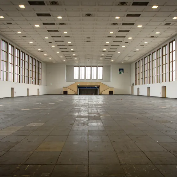 Große, leere Halle mit vielen Fenstern und hoher Decke, Tageslicht fällt herein.