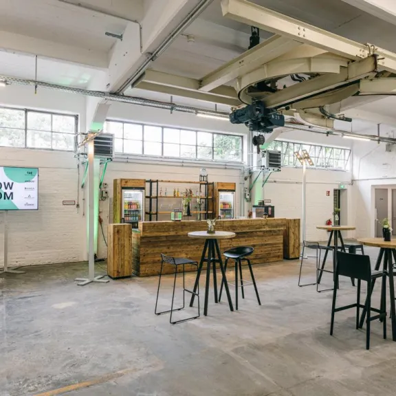 Heller, moderner Innenraum mit Bar, Stehtischen und Hockern in einer Industriehalle.