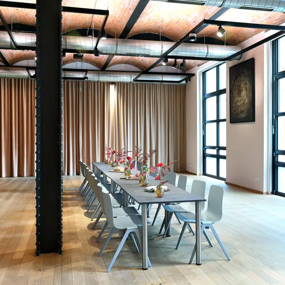 Großer, heller Raum mit langem gedecktem Tisch und modernen Stühlen, Holzfußboden, loftartig.