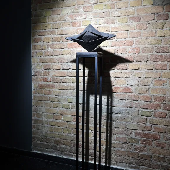 Schwarze Skulptur auf hohem Metallgestell vor beleuchteter Backsteinwand.