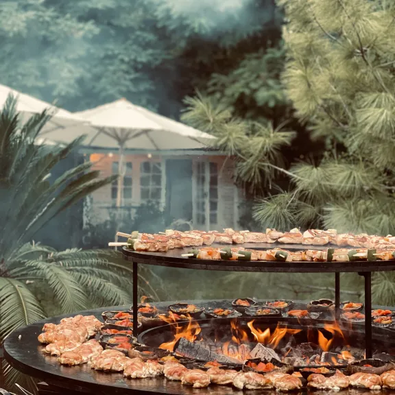 Grill mit Fleischspießen im Garten, Rauch steigt auf, im Hintergrund Bäume und Sonnenschirme.