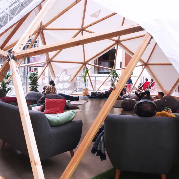 Menschen entspannen auf Sofas in einem offenen, modernen Holzzelt in einer Halle.