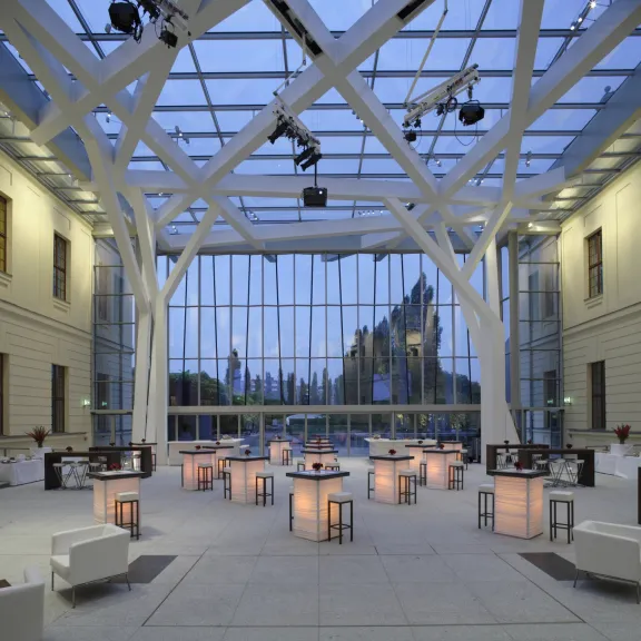 Modernes Atrium mit Glasdach, weißen Sitzmöbeln und beleuchteten Stehtischen.
