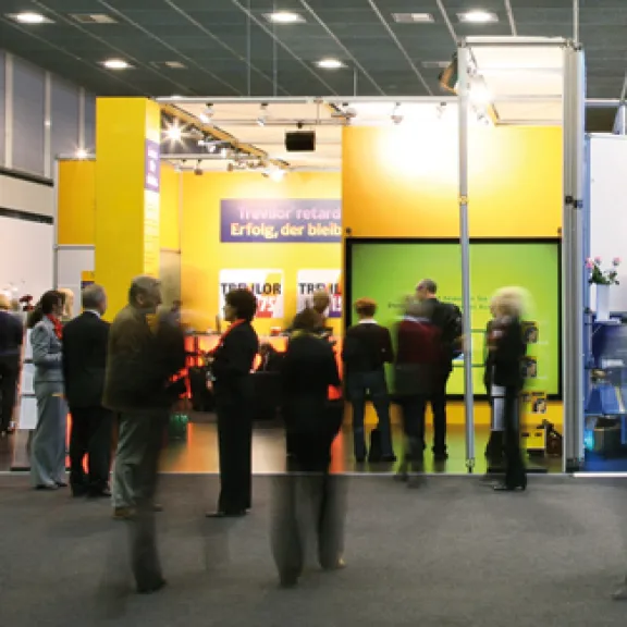 Menschen stehen und gehen vor einem gelb beleuchteten Messestand.