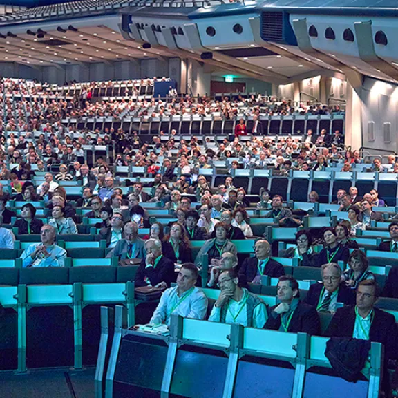 Großer Konferenzsaal mit vielen sitzenden, aufmerksam lauschenden Menschen.