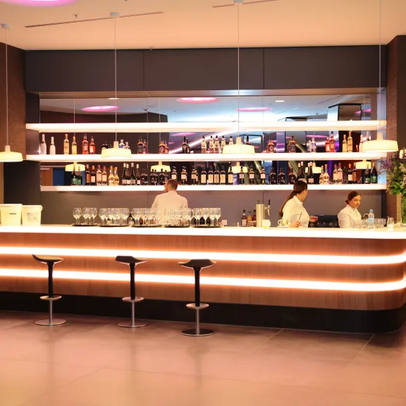 Moderne Bar mit drei Barhockern, zwei Barkeepern und beleuchteten Regalen im Hintergrund.