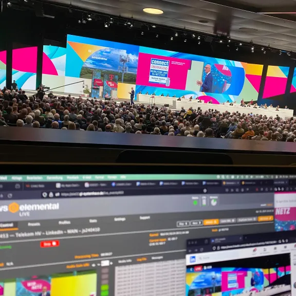 Ein großes Publikum verfolgt die Hauptversammlung der Deutschen Telekom. Im Vordergrund ist ein Bildschirm zu sehen, der den Cloud-Encoder und den LinkedIn-Stream für die Veranstaltung überwacht.
