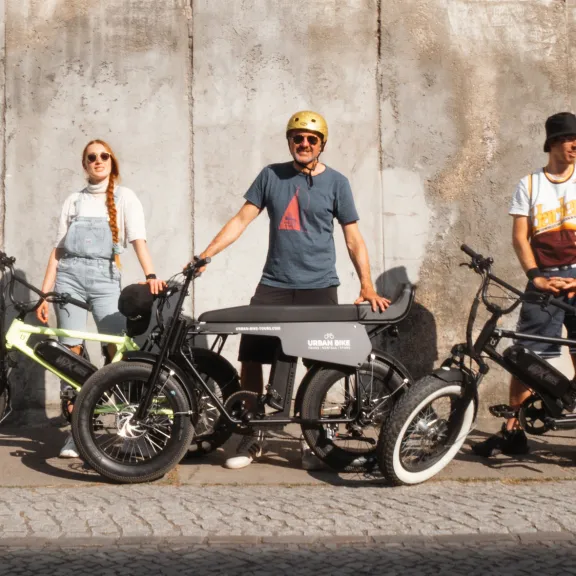 Drei Personen mit E-Bikes stehen vor einer Betonwand in der Sonne.