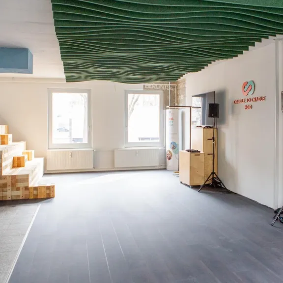 Heller Veranstaltungsraum mit großen Fenstern, modularer Holztribüne und flexibler Technik. An der Decke wellenförmige Elemente, an der Wand das Logo der Cradle to Cradle NGO. Offene Fläche für Workshops und Events.