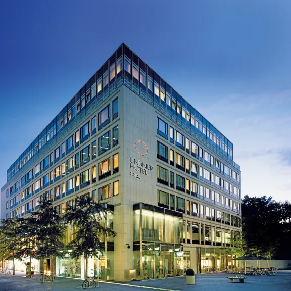 Modernes Hotelgebäude bei Dämmerung mit beleuchteten Fenstern und Bäumen davor.