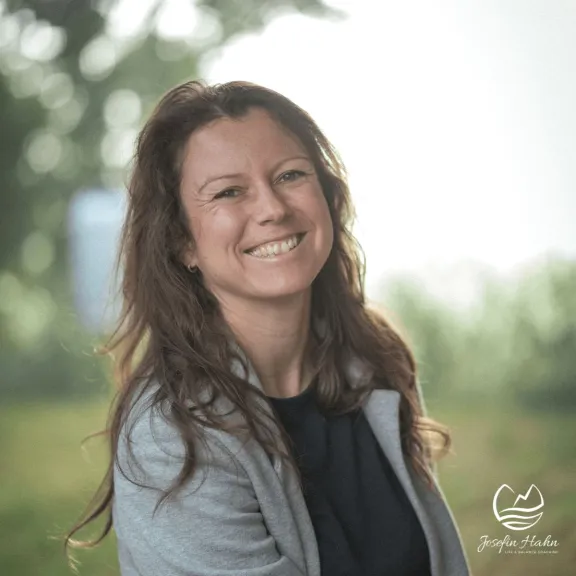 Lächelnde Frau mit langen braunen Haaren im Freien, unscharfer grüner Hintergrund.