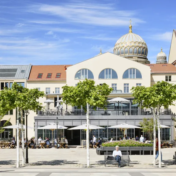 Belebter Platz mit Bäumen und Café vor historischem Gebäude mit Kuppel.